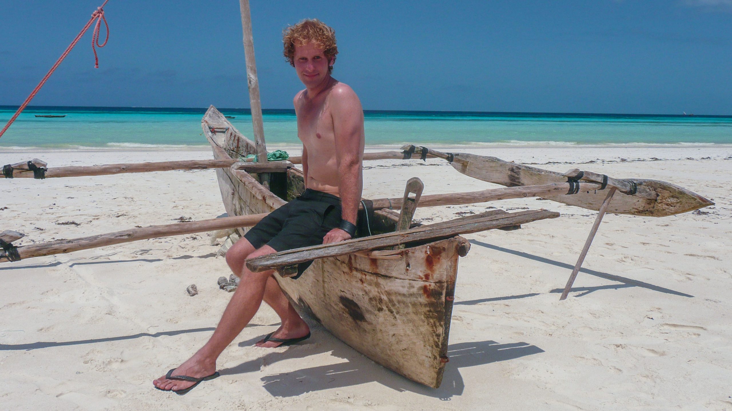 Shirtless Ben sitting on dugout canoe on beach of Zanzibar Tanzania